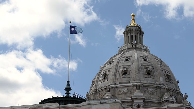A state capitol building with a domed roof stands behind mature trees on a clear day.