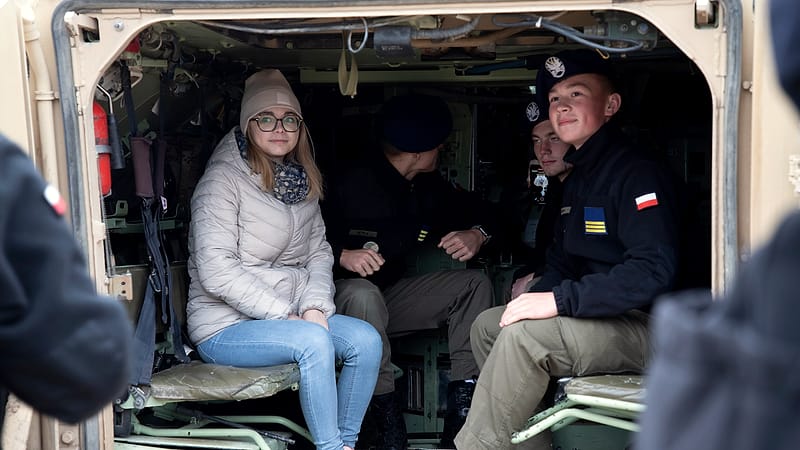 A uniformed service member explains equipment to a group of visiting students beside an armored vehicle.