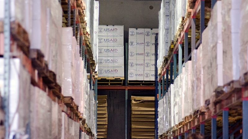 A forklift stands beside stacked pallets in a warehouse aisle with safety markings on the floor.
