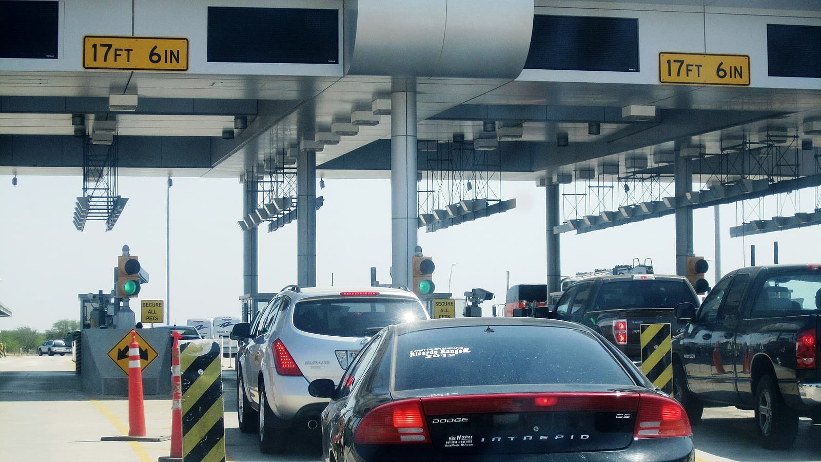 Vehicle lanes queue toward a covered land border inspection plaza with overhead signage and booths.