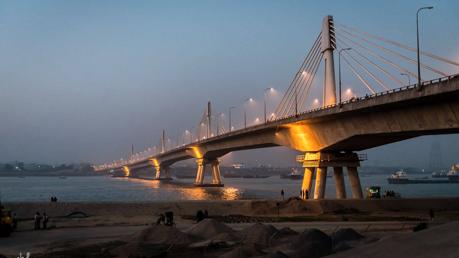 A long road bridge photographed from the bank, carrying traffic across a wide river toward a distant city.