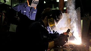 A worker in protective gear welds a metal joint in a ship's workshop while sparks fall around the bench.